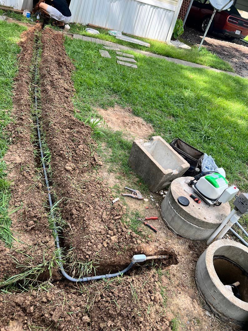 A person in a grassy area digs a trench for a black pipe near a concrete structure.