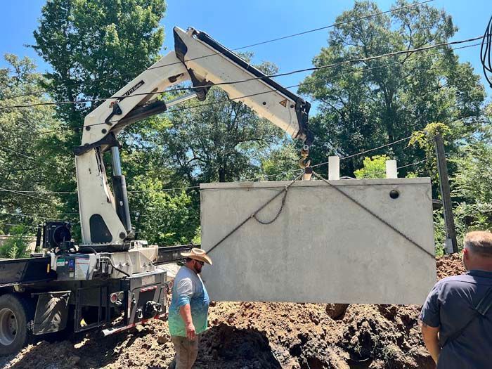 Crane lifting a concrete septic tank. Two men watch installation outdoors. Sunny day.