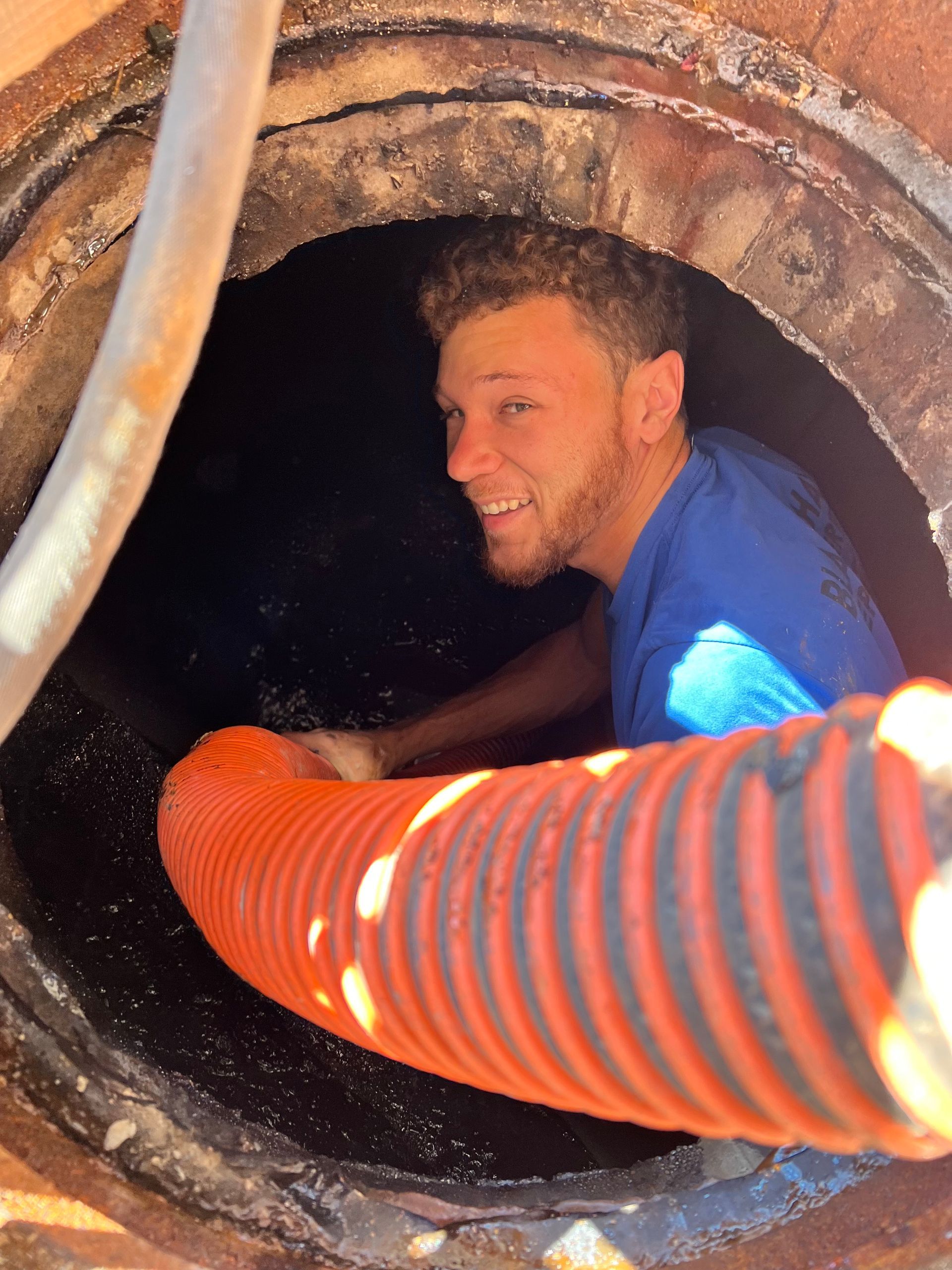 Man smiles while inside a dark, circular hole; an orange hose is extended towards him.