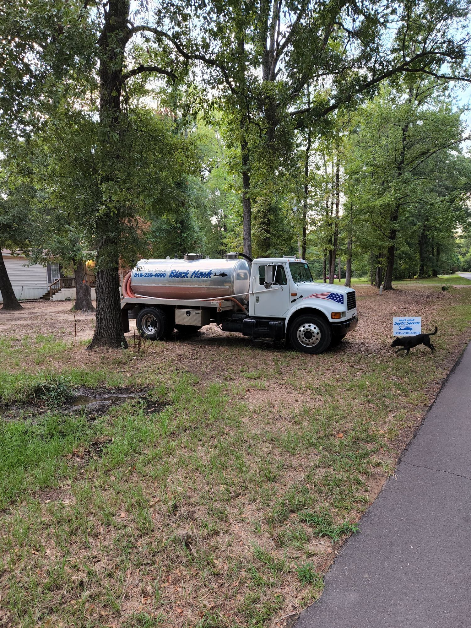 A white water truck parked in a grassy area near trees. A dog sits on the right.