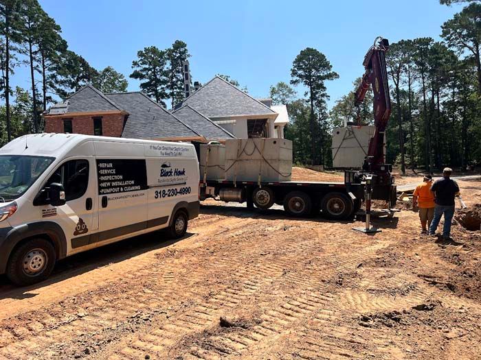 A septic tank on a flatbed being lifted by a crane at a construction site. Men standing nearby.