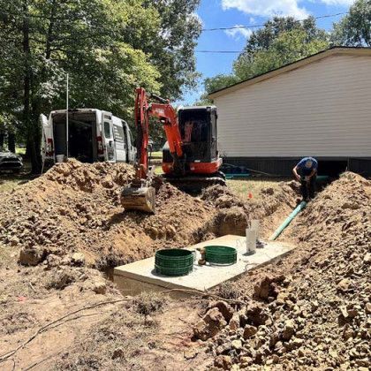 Excavator installing a septic tank near a house. Man working on pipes. Dirt mound, sunny day.
