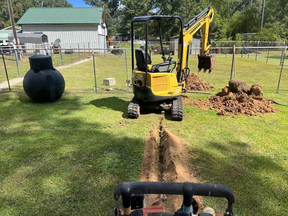A yellow excavator digs a trench in a grassy yard, near a septic tank and fence.