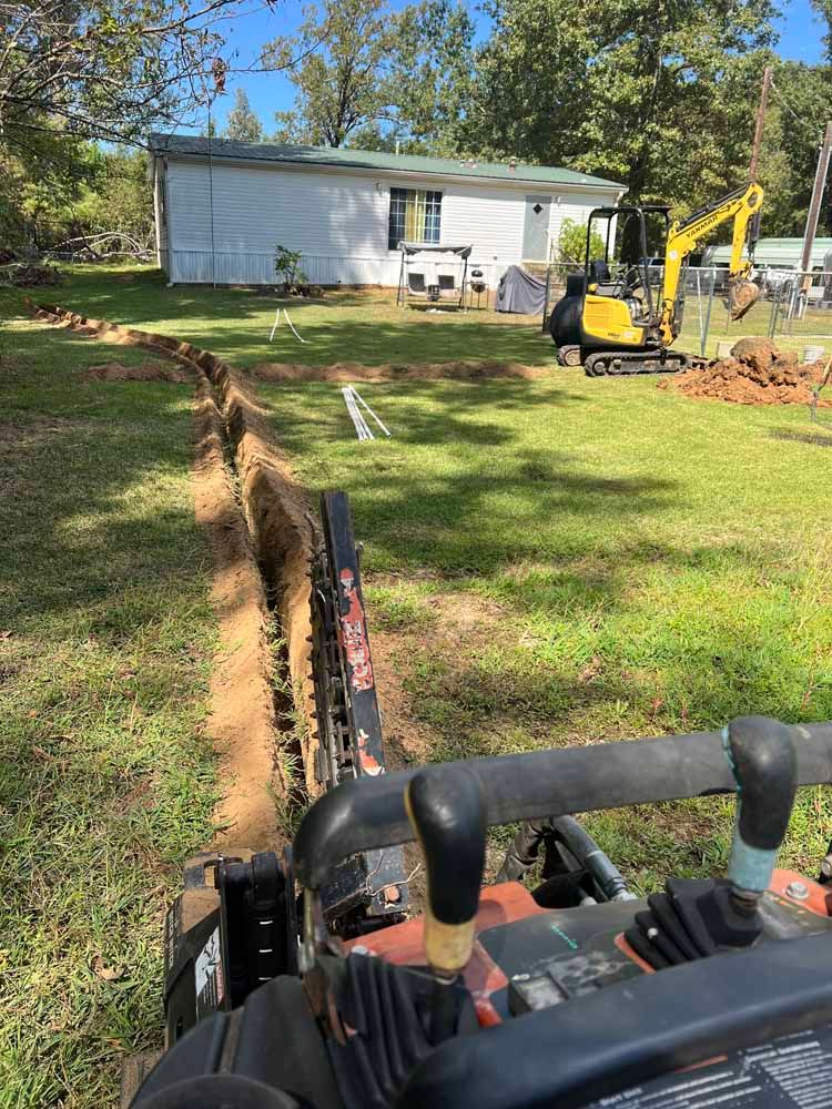A trench is being dug in a grassy yard near a small house with an excavator.