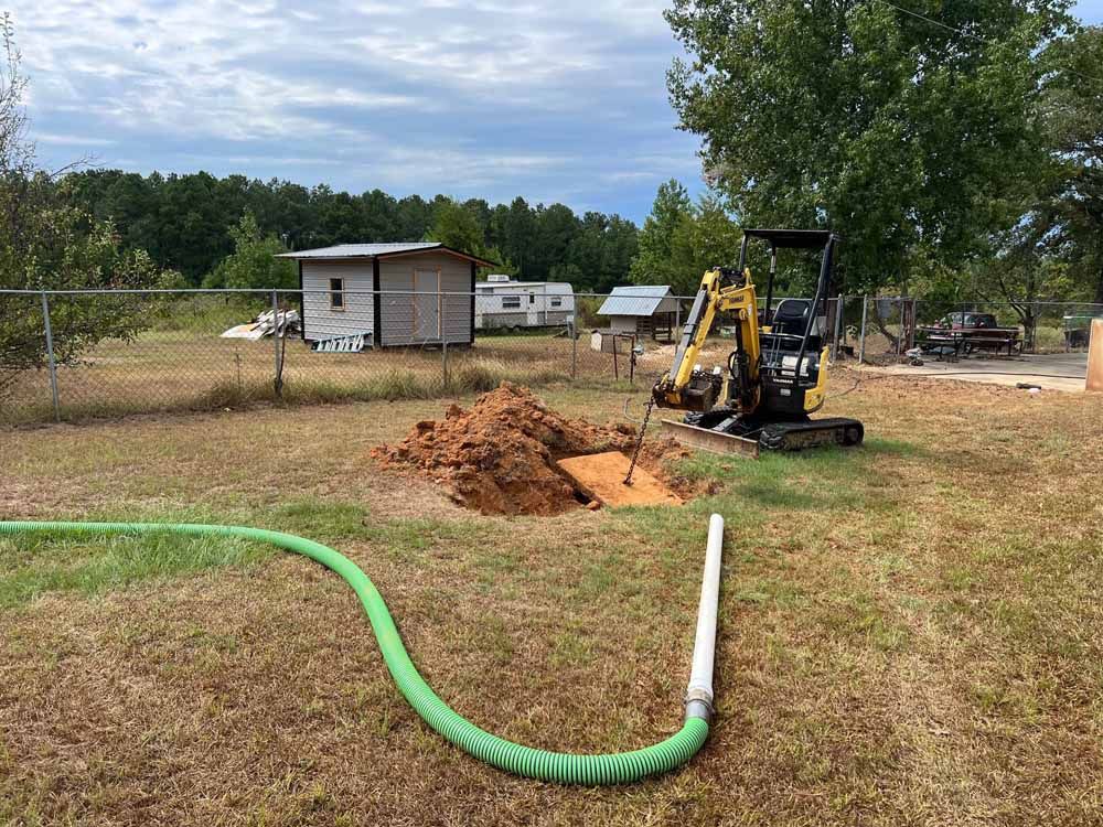 Mini excavator digging hole in yard, green hose nearby. Small buildings, trees, and cloudy sky in background.
