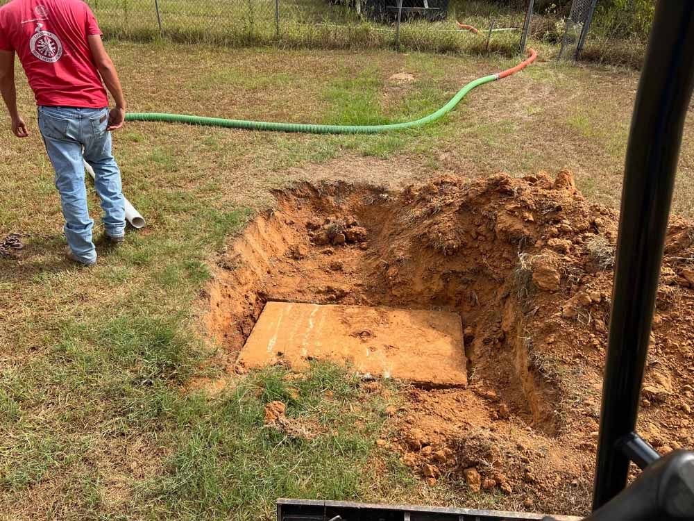 Man in red shirt near a square hole in the ground with a concrete cover, a green hose, and a fence.