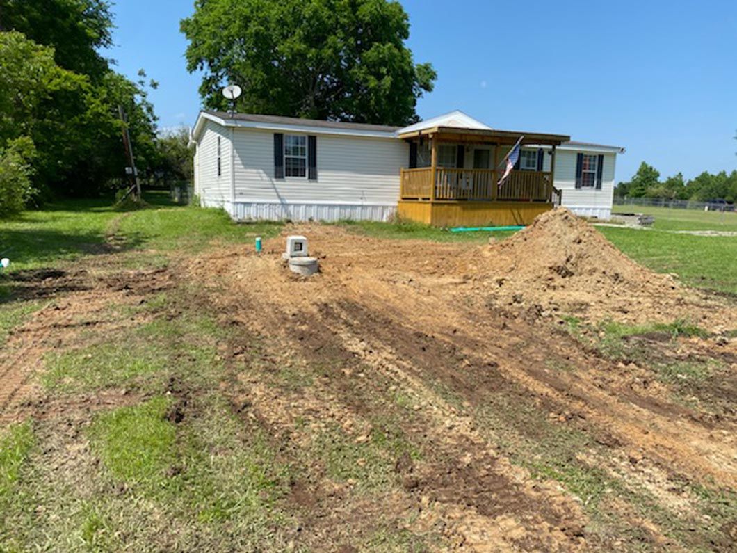 Mobile home with porch, dirt piles, and construction in a grassy yard under a blue sky.