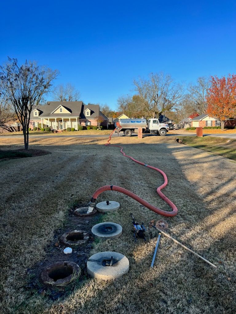 A septic tank being pumped. A truck with a hose in a yard. Residential houses in the background.