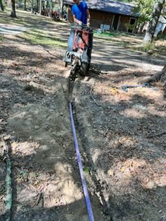 Man operating trenching machine, laying purple cable in a yard with trees.