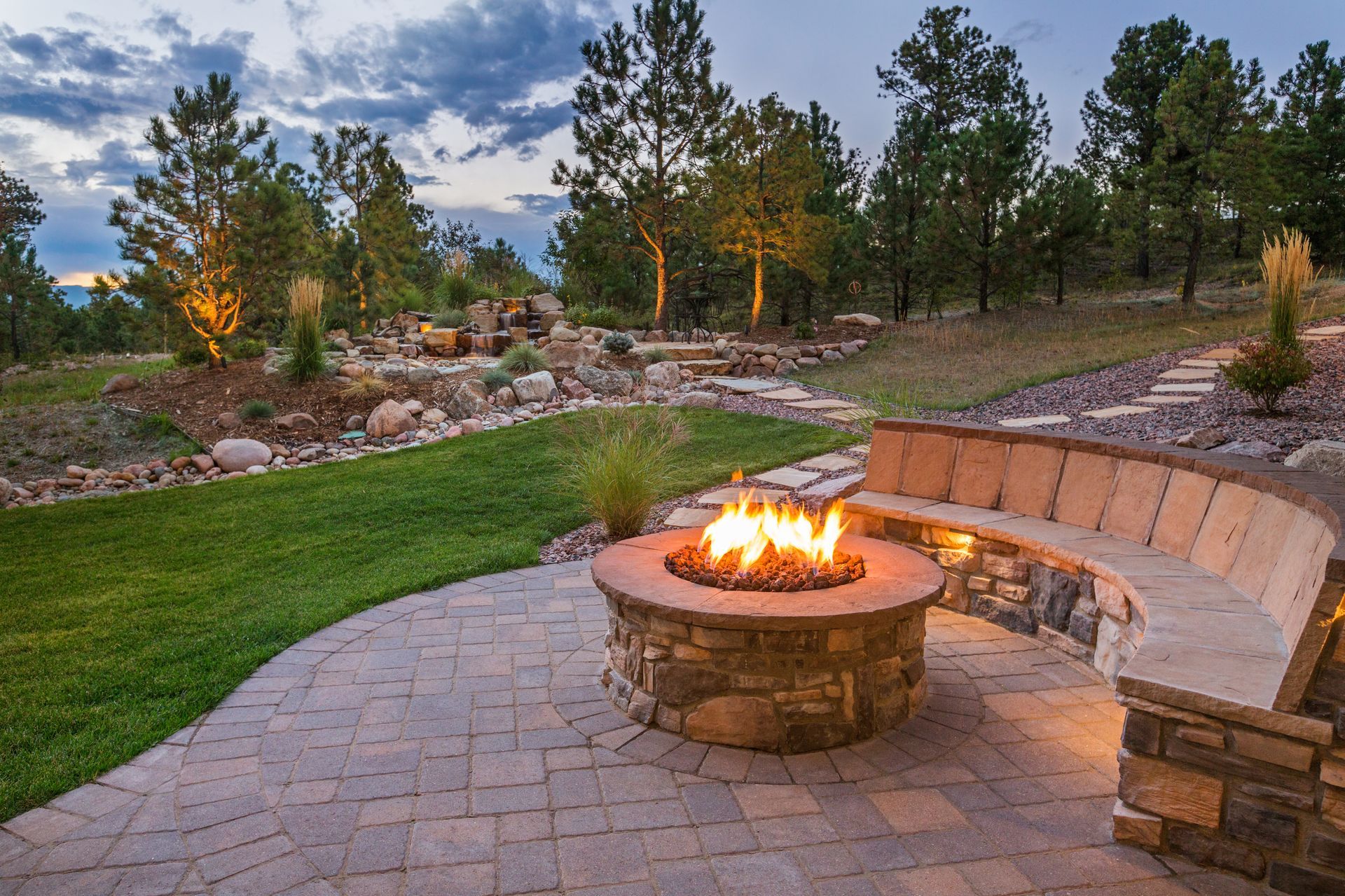 Outdoor stone fire pit with curved seating on a paved patio, set in a landscaped backyard. Outdoor stone fire pit with curved seating on a paved patio, set in a landscaped backyard.