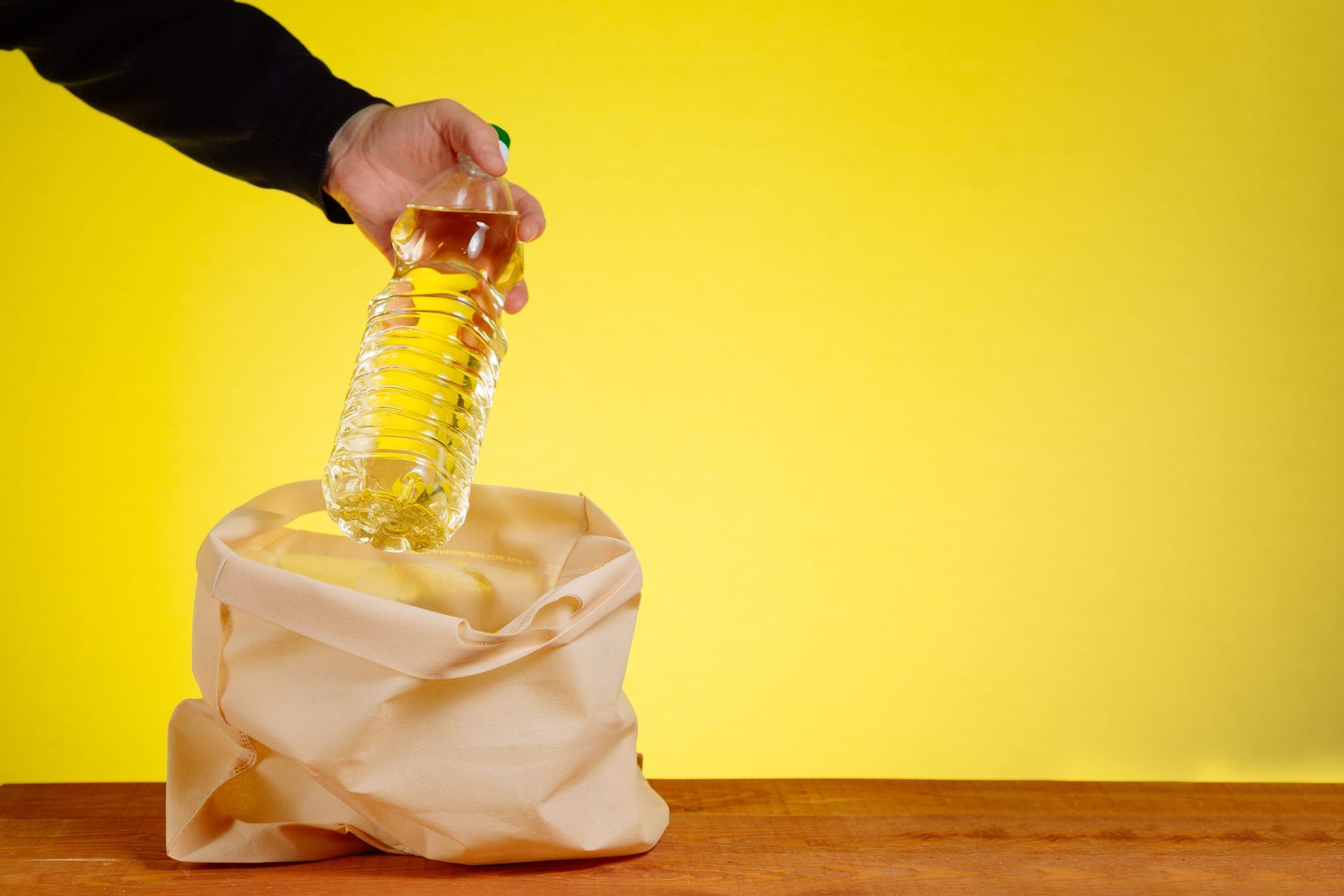 A hand holds a plastic bottle of golden oil over an open beige cloth bag against a bright yellow background.