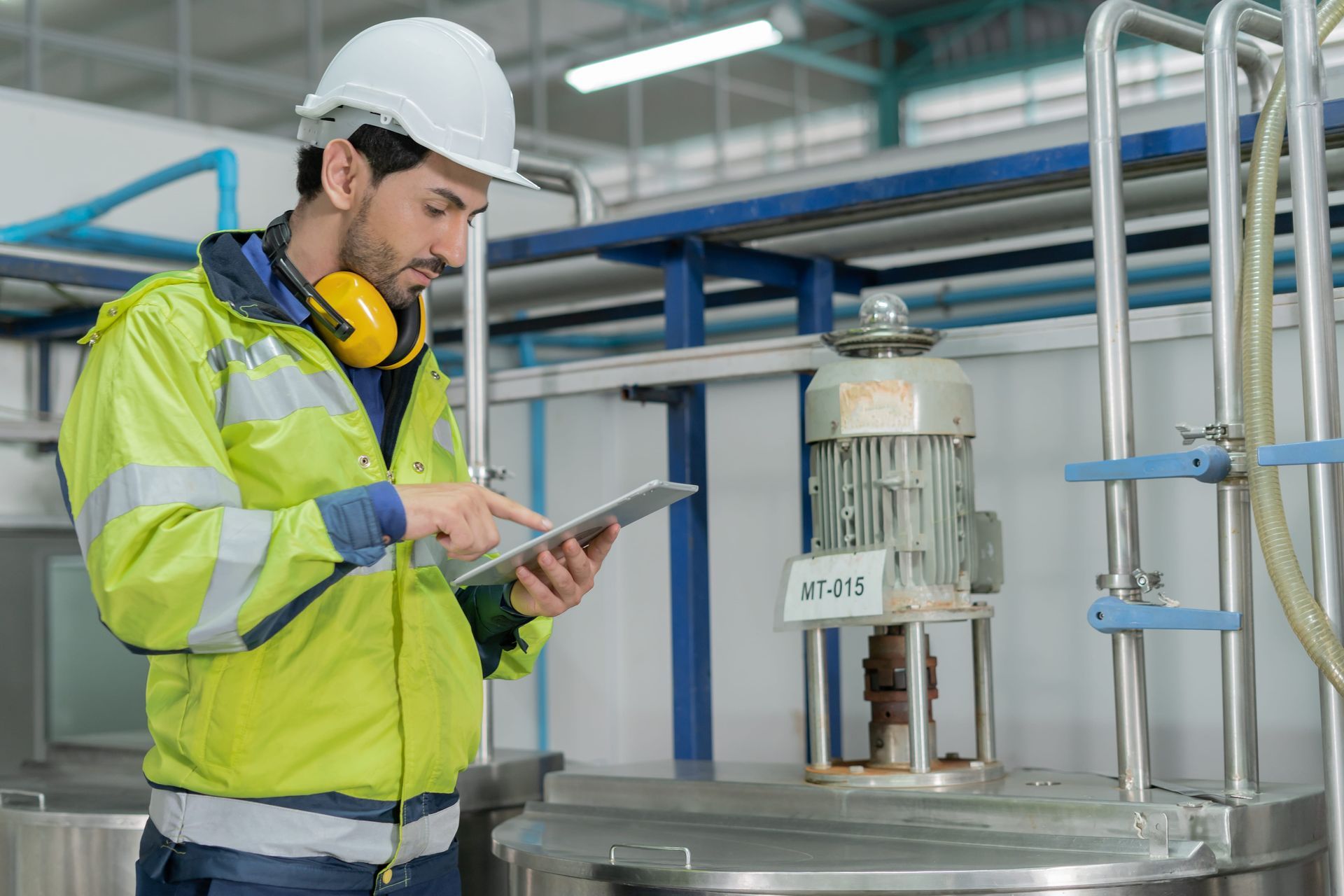 A person in a high-visibility jacket and hard hat uses a tablet to inspect industrial machinery in a factory.