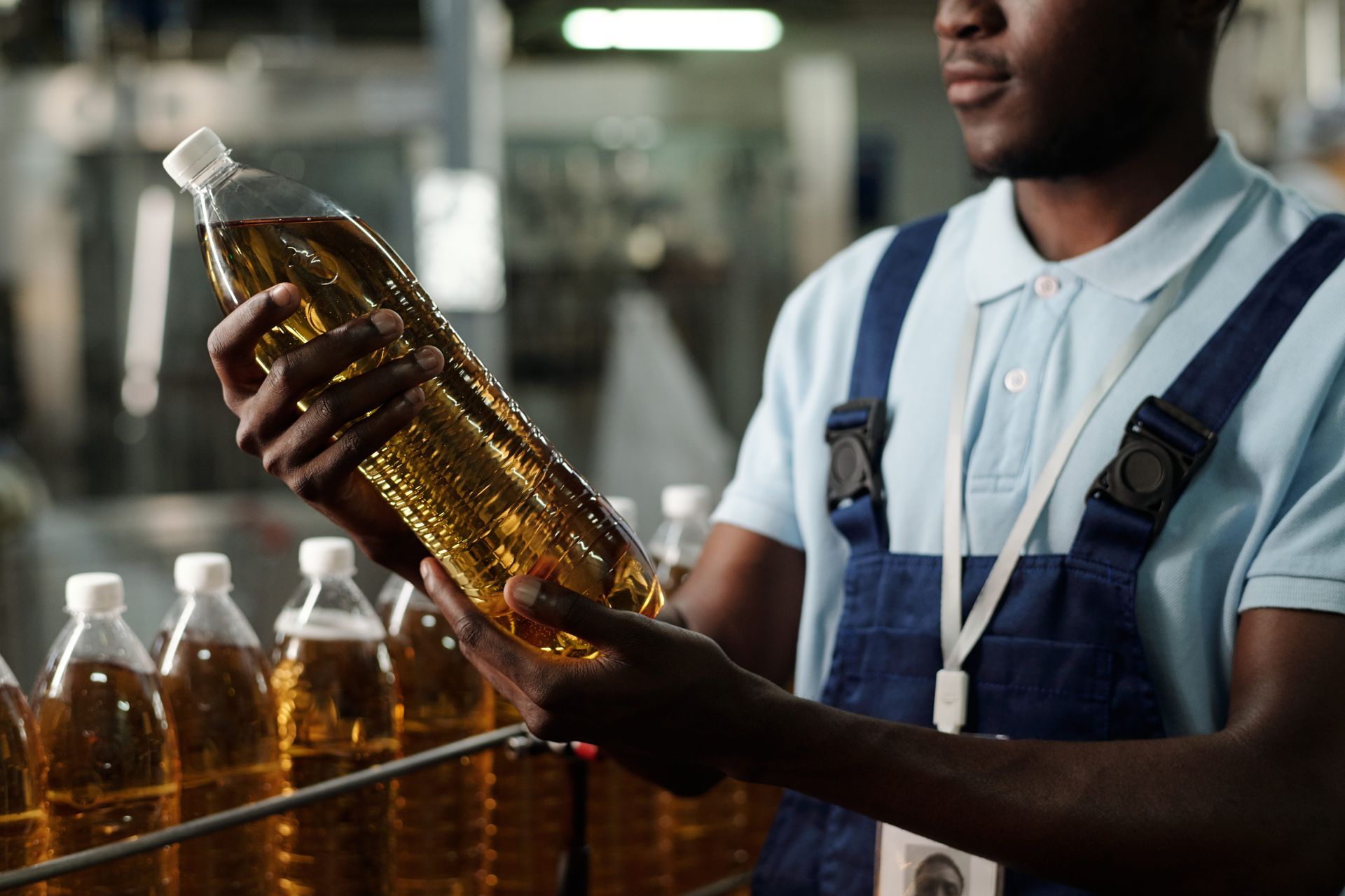 A worker in blue workwear inspects a bottle of golden liquid on a factory production line.