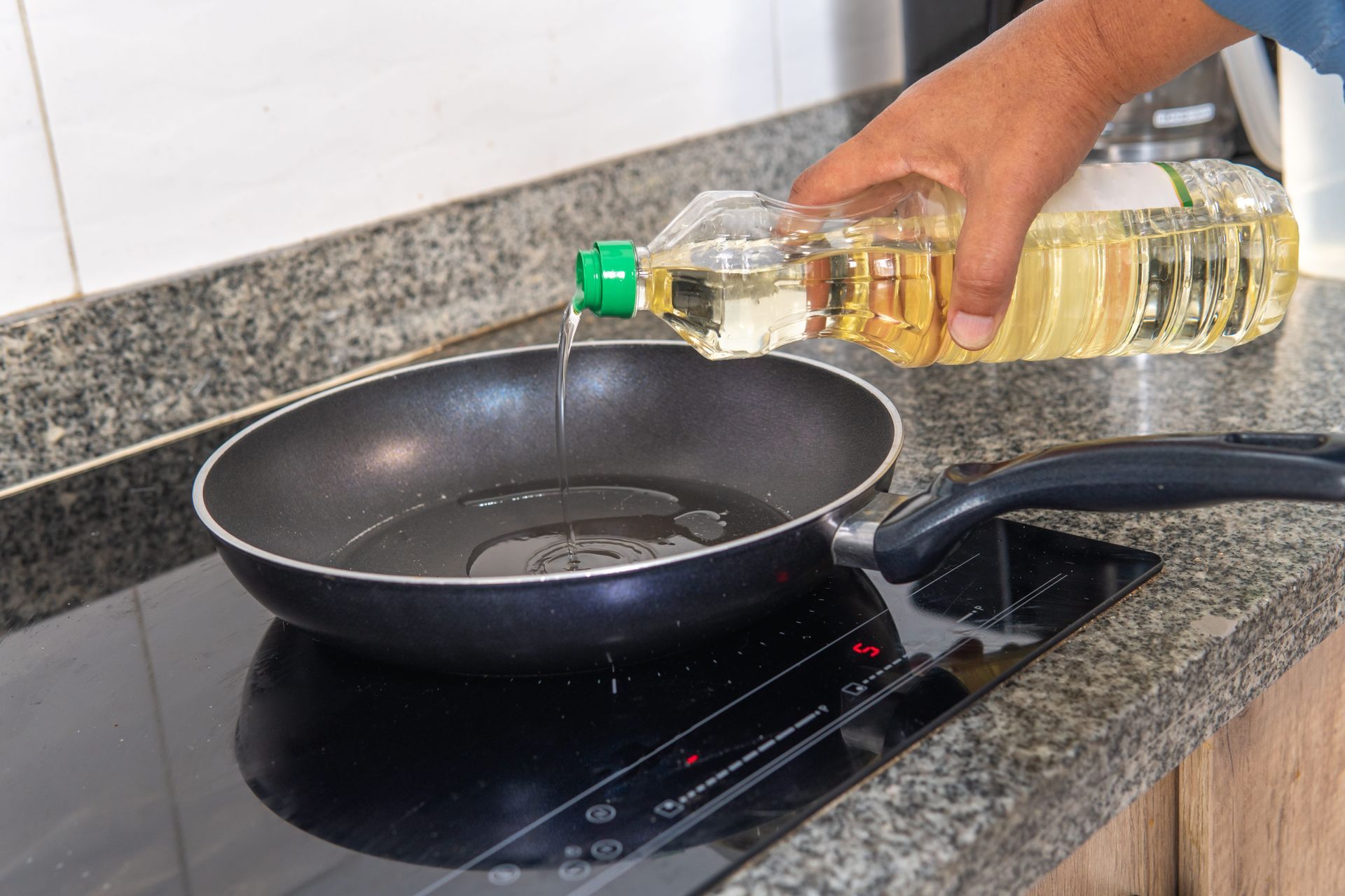 A person pours cooking oil from a plastic bottle into a nonstick frying pan on a black induction stovetop.