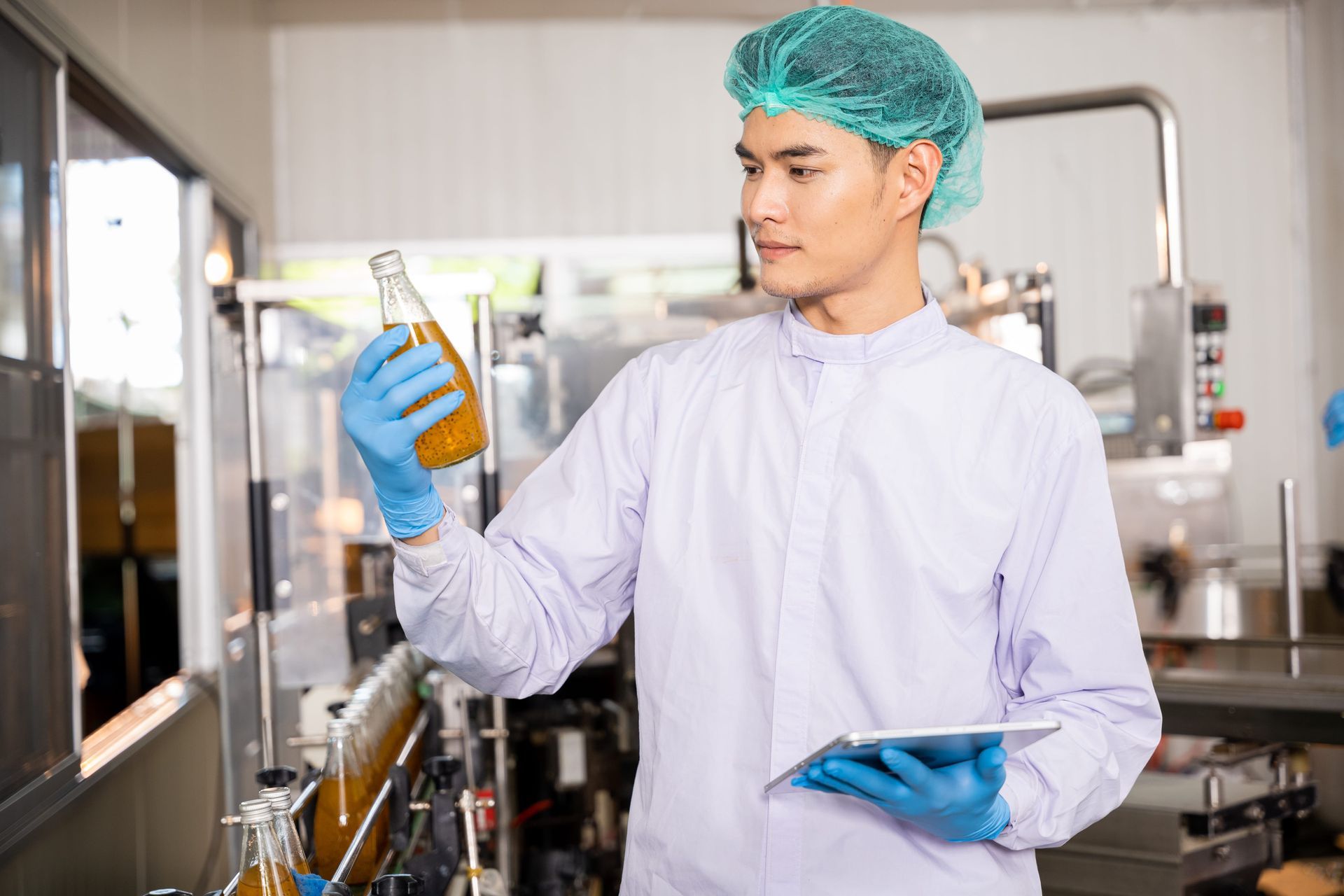 A worker in a food processing plant wears a hairnet and gloves, inspecting a glass bottle of liquid while holding a tablet.