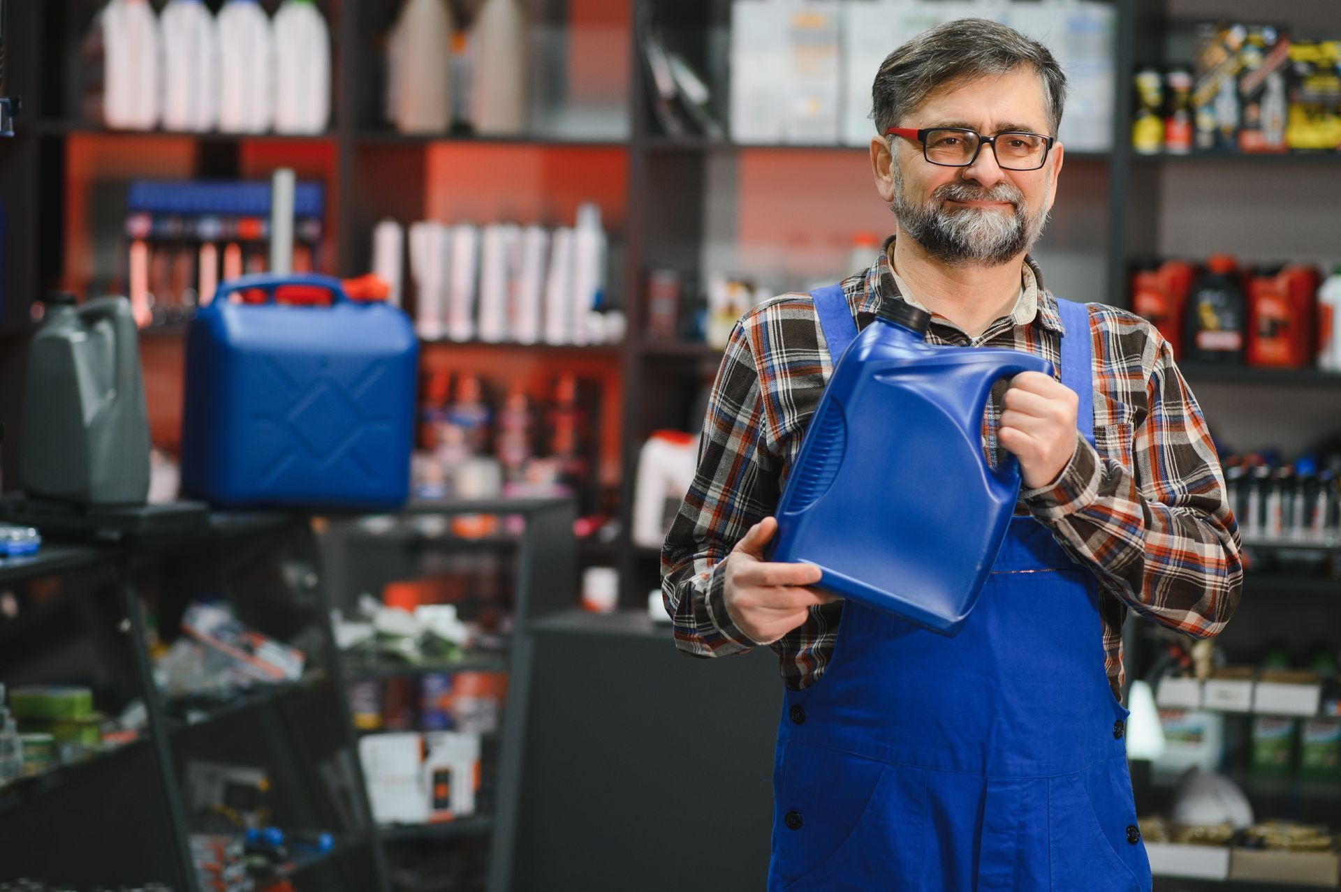 A worker in blue coveralls holding a blue plastic jug inside an automotive supply store with shelves of products behind him.