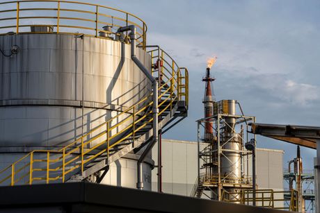 Large silver industrial storage tanks and a processing tower with a burning flare under a cloudy sky.