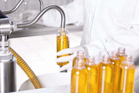 A person in a white lab coat and gloves fills a clear bottle with golden liquid from a metallic industrial nozzle.