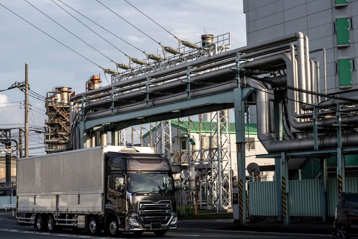 A dark truck with a white trailer drives under a large metal industrial pipe structure in an urban setting.
