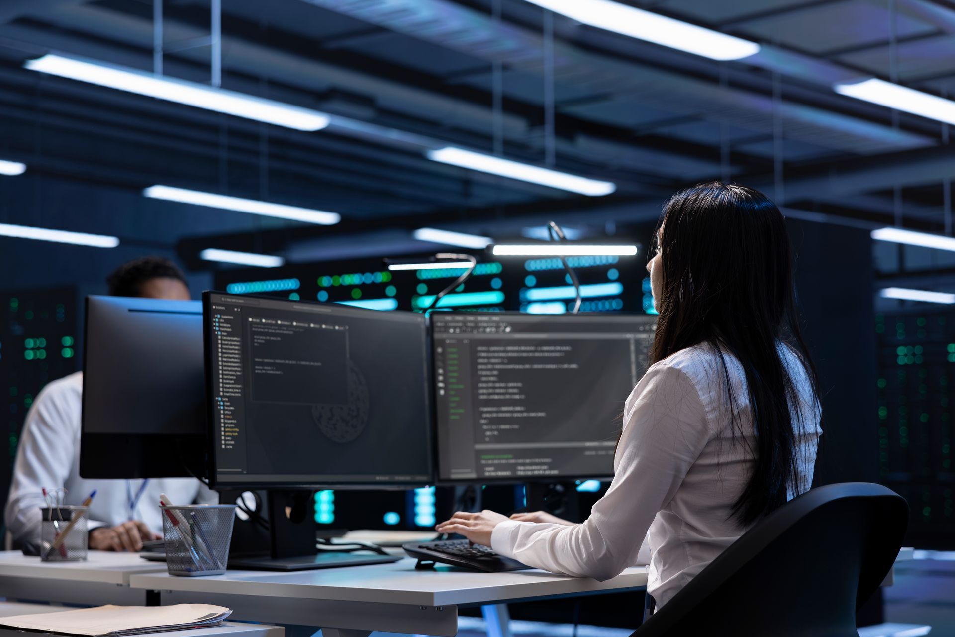 Professionals work at desks in a dimly lit, high-tech server room, viewed from behind as they monitor code on dual screens.
