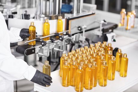 A worker in a white coat and black gloves places amber-colored plastic bottles onto a factory labeling machine.
