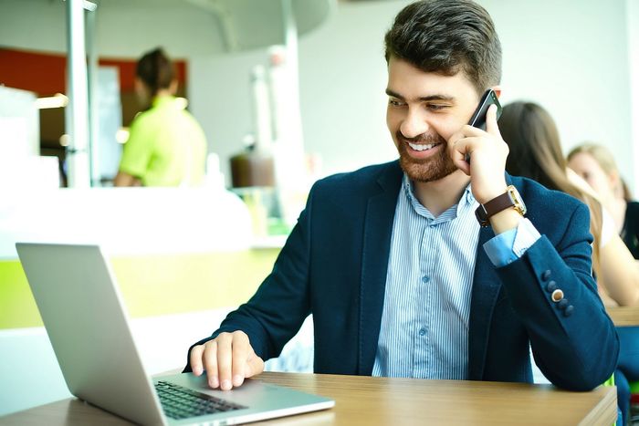 A professional in a blazer sits at a table using a laptop while talking on a smartphone in a bright, modern cafe.