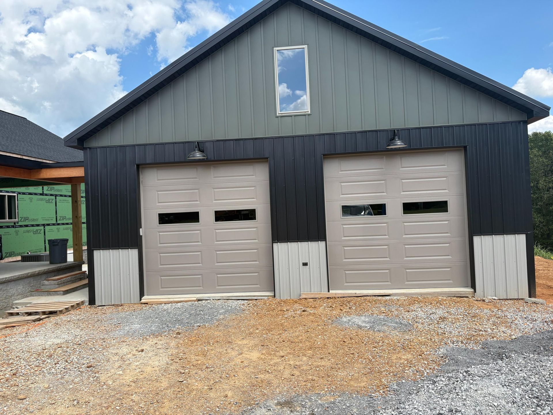 Two-car garage with gray-beige doors and black and gray siding. Gravel driveway, blue sky reflection in window.