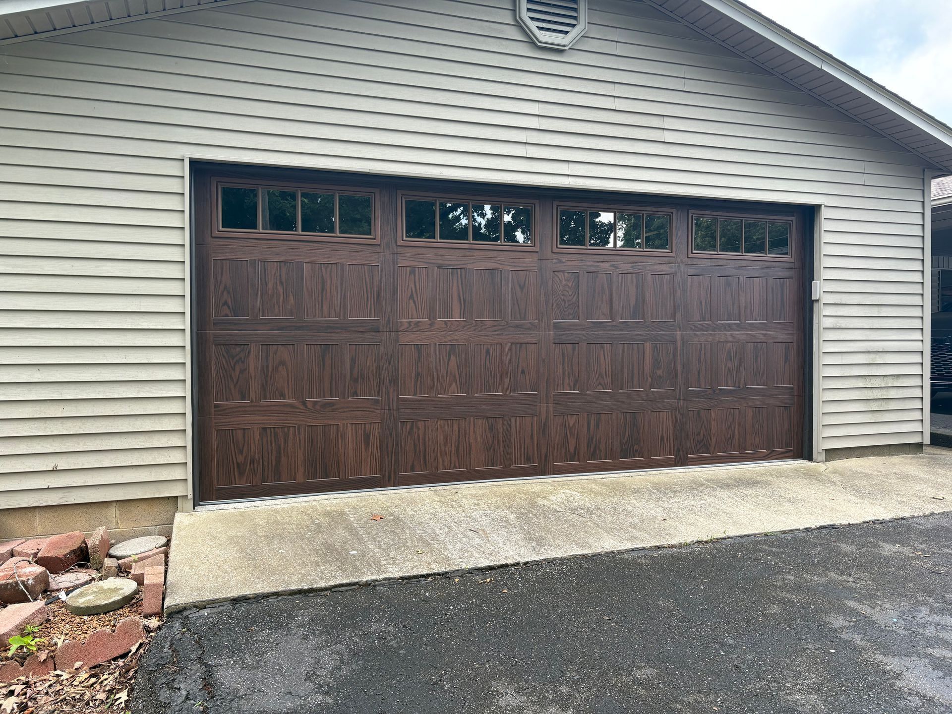 Brown garage door on a tan house, with glass paneling at top. Concrete driveway and asphalt.