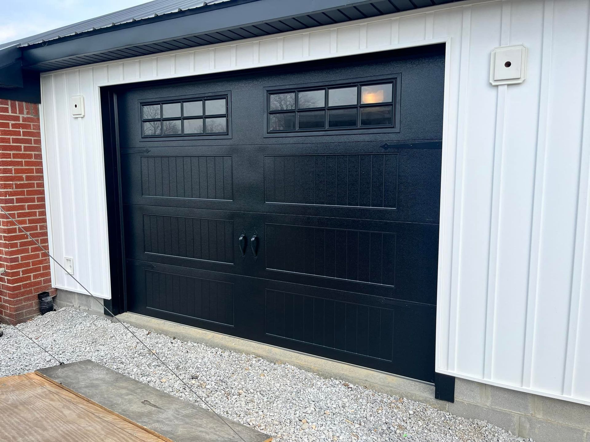 Black garage door with windows on a white building; gravel base.
