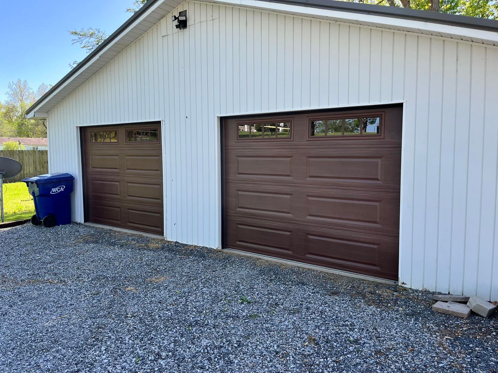 Two-car garage with brown doors and white siding, set on a gravel driveway with a blue trash can.