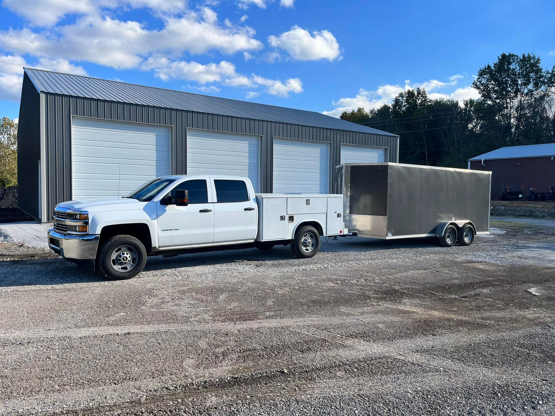 White truck with a trailer in front of a metal building on a gravel lot.