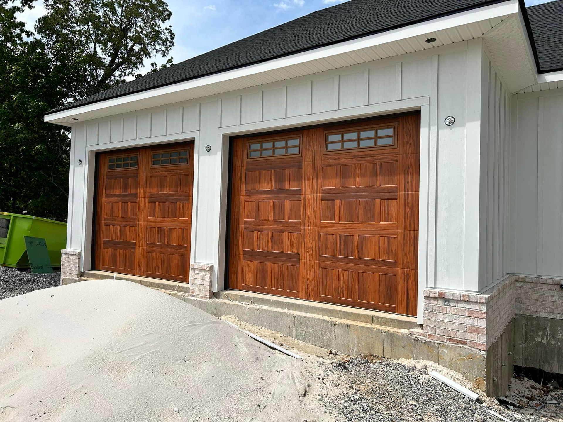 Two brown wooden garage doors in a white-paneled building with stone accents, gravel, and sand in front.