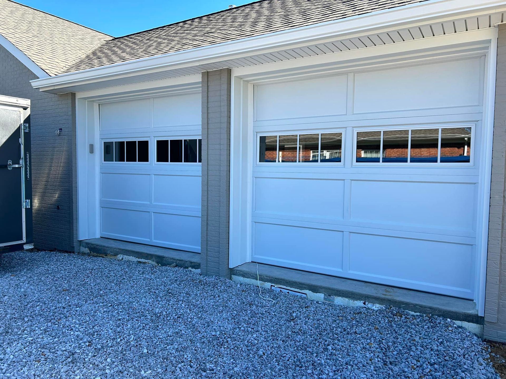 Two white garage doors with windows, set in a white frame, gray gravel, and white trim.