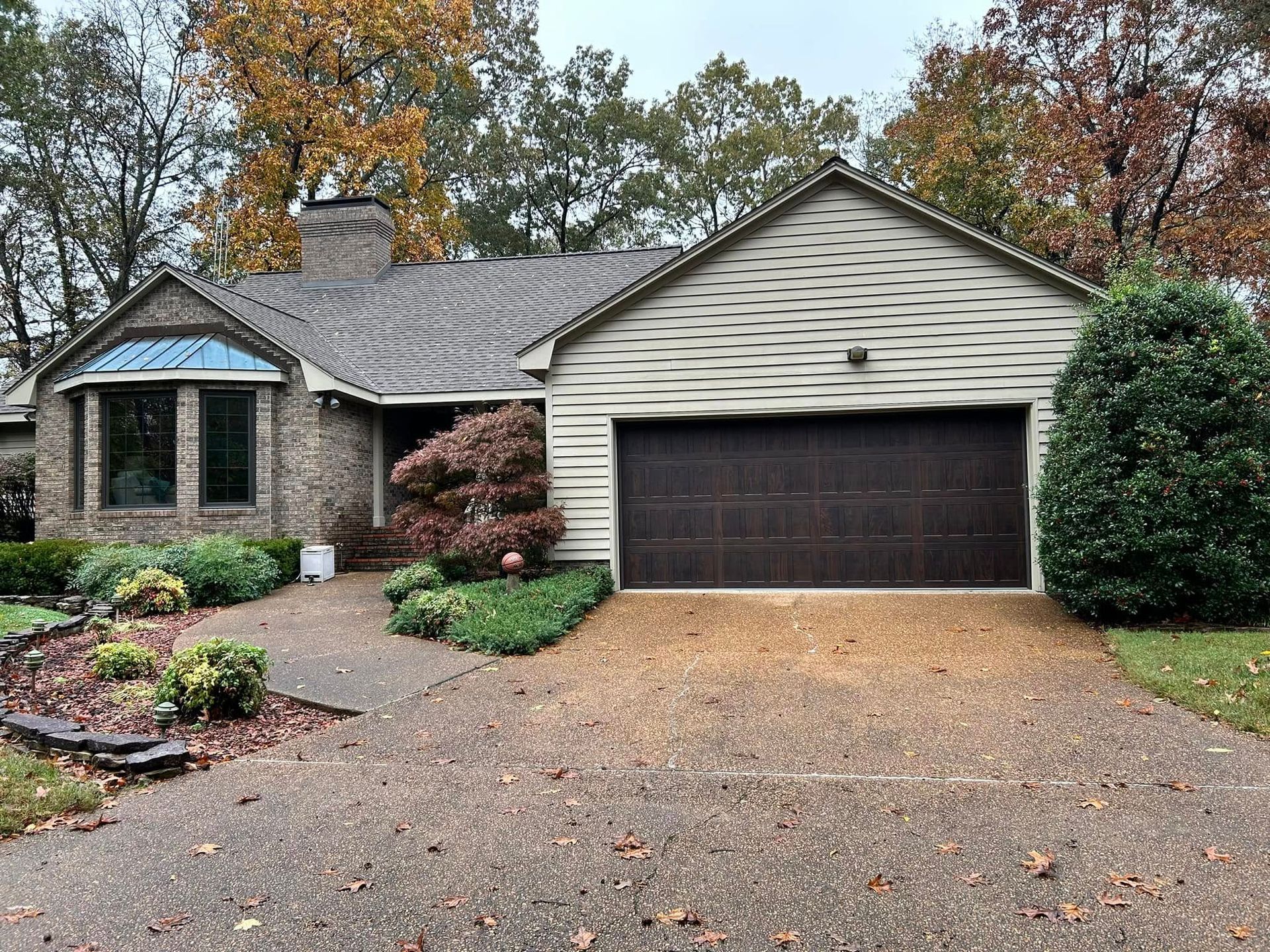 One-story brick house with brown garage door, driveway covered in leaves, surrounded by trees.