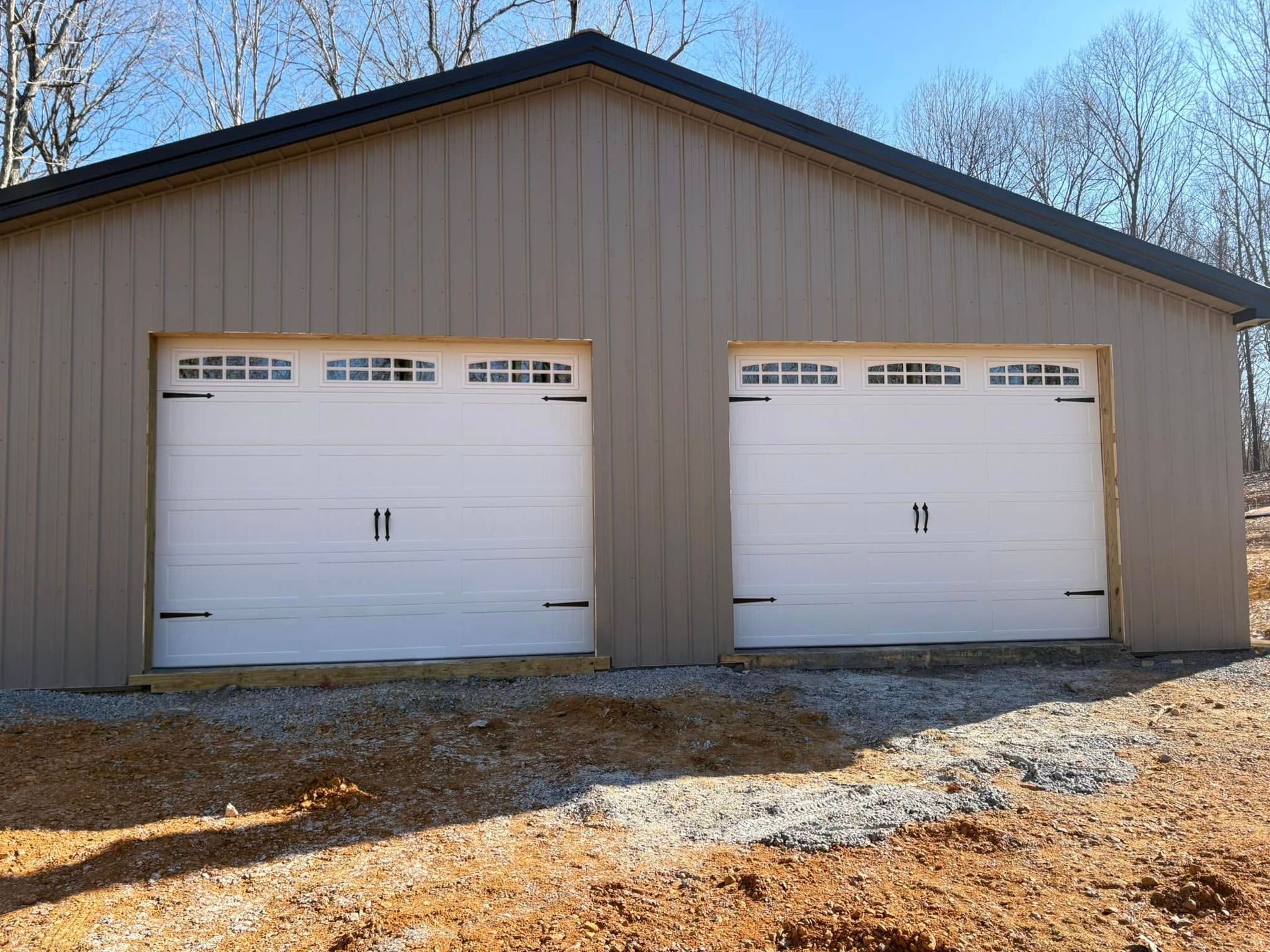 Tan garage with two white garage doors, set on gravel and dirt under a bright sky.