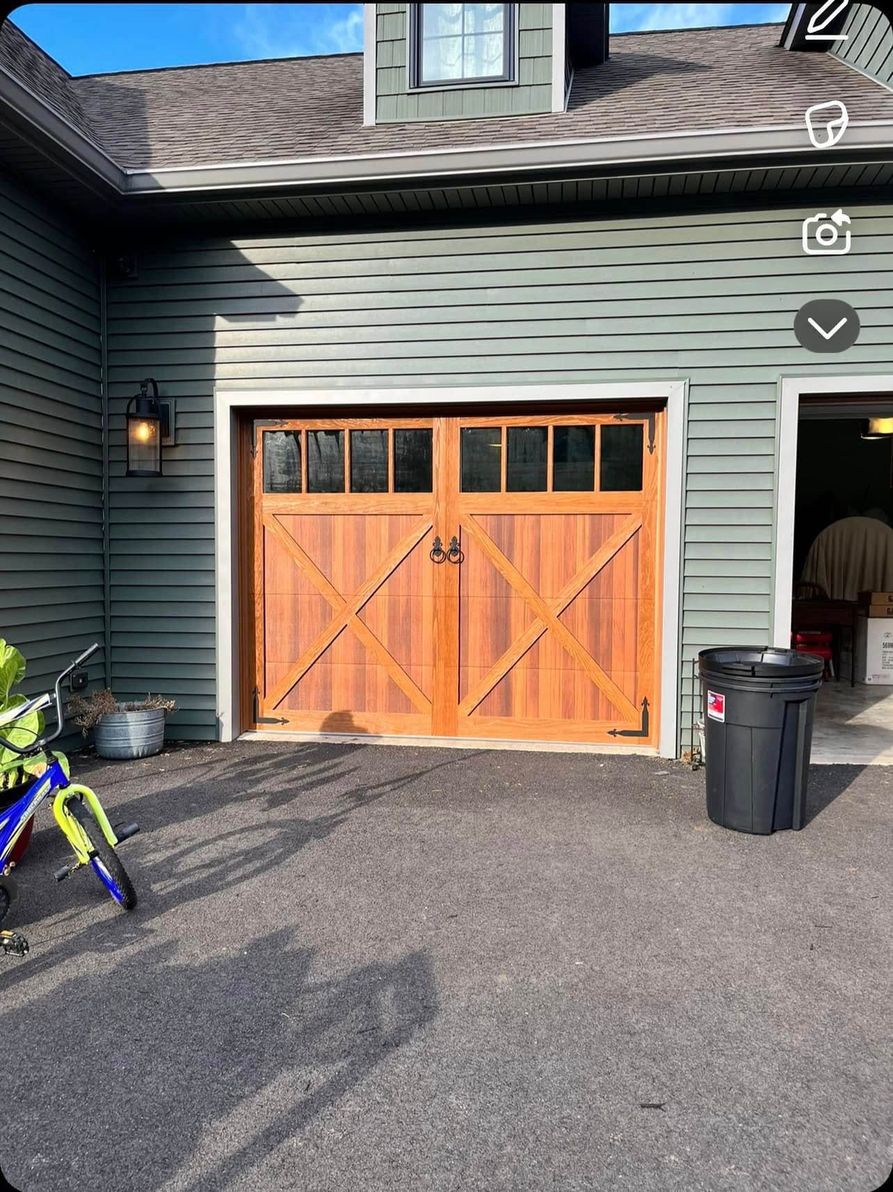 Wooden garage doors with an X-design, surrounded by green siding. A bike and trash can sit nearby.