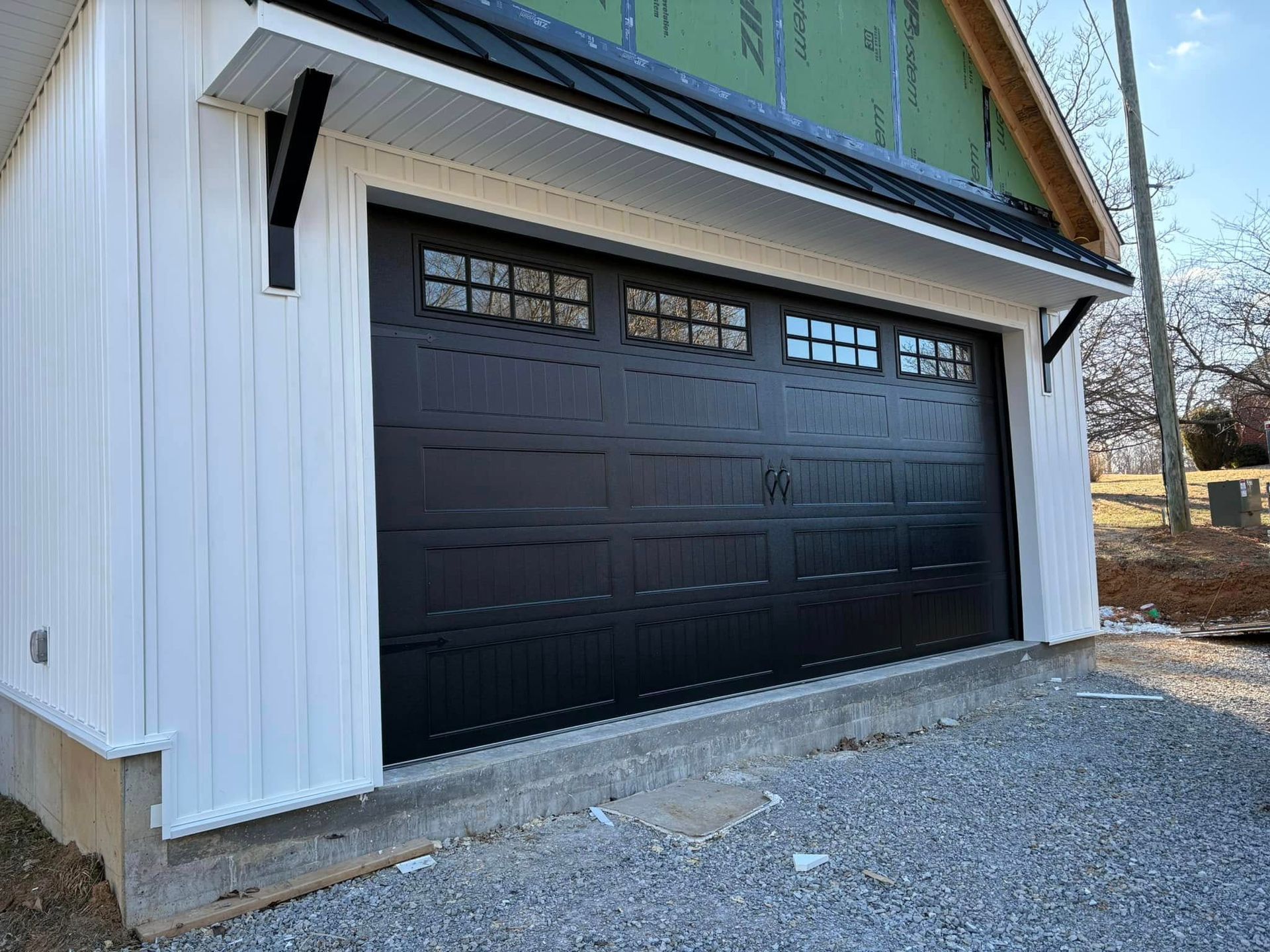 Black garage door with small windows, white siding, black accents, and gravel driveway.