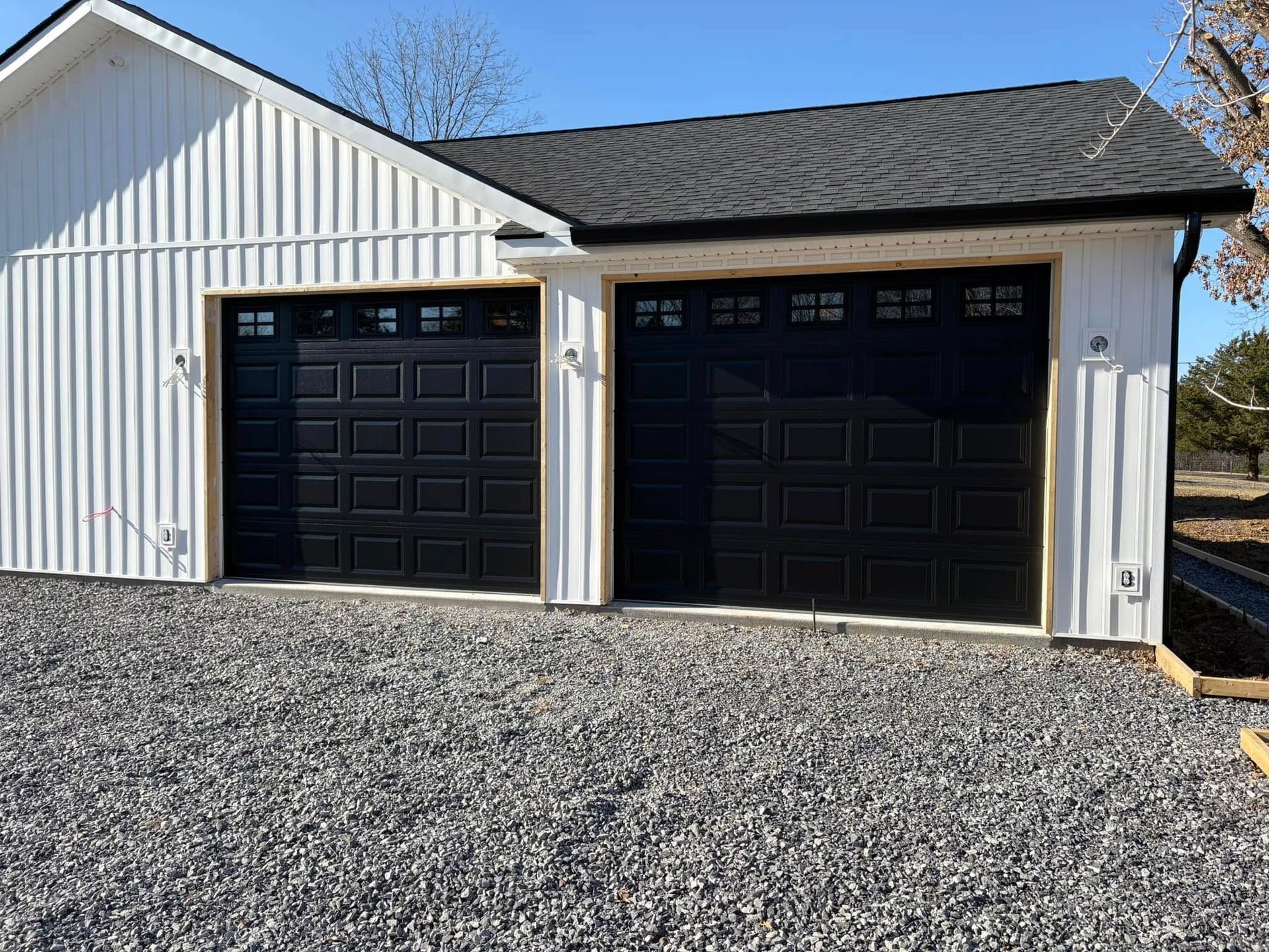 Two black garage doors on a white building with a dark roof, set on a gravel driveway.