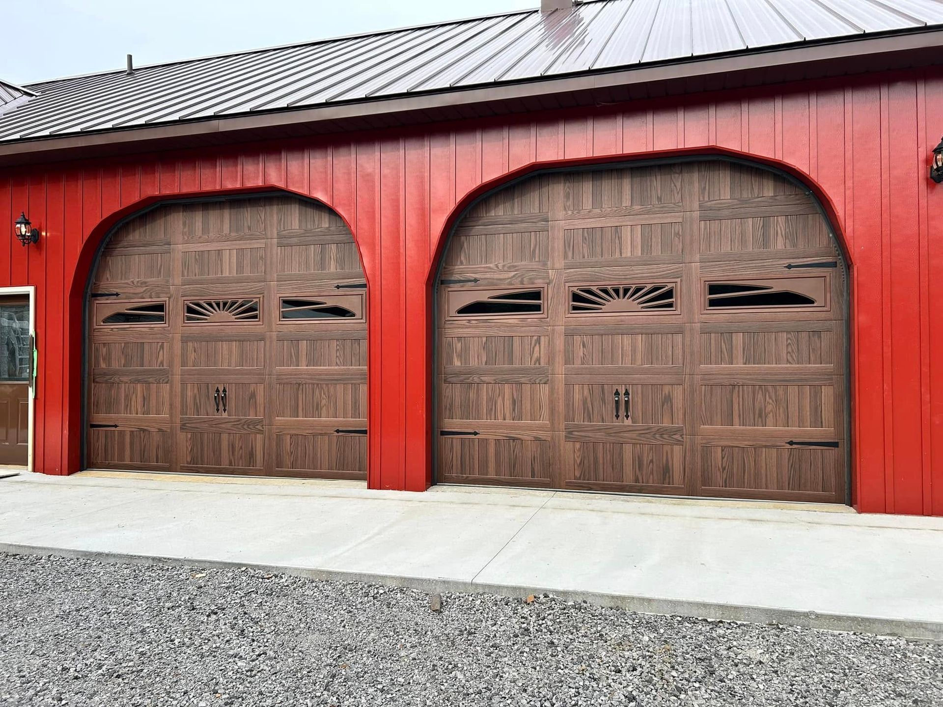 Red barn with two arched wooden garage doors, brown roof, gravel driveway.