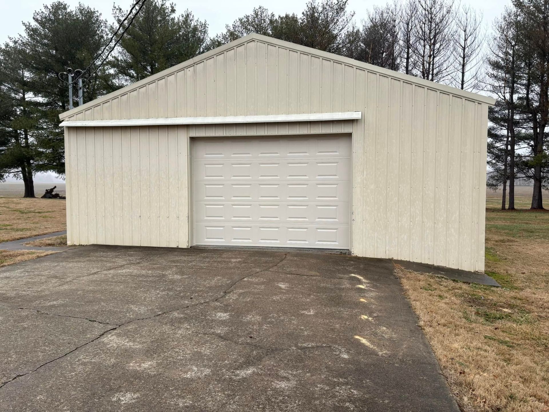 A beige metal garage with a closed door sits on a cracked asphalt driveway.