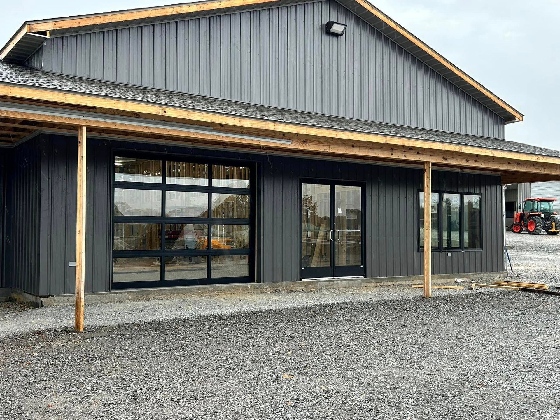 Gray building with glass garage door and windows under a wooden overhang. Gravel ground.