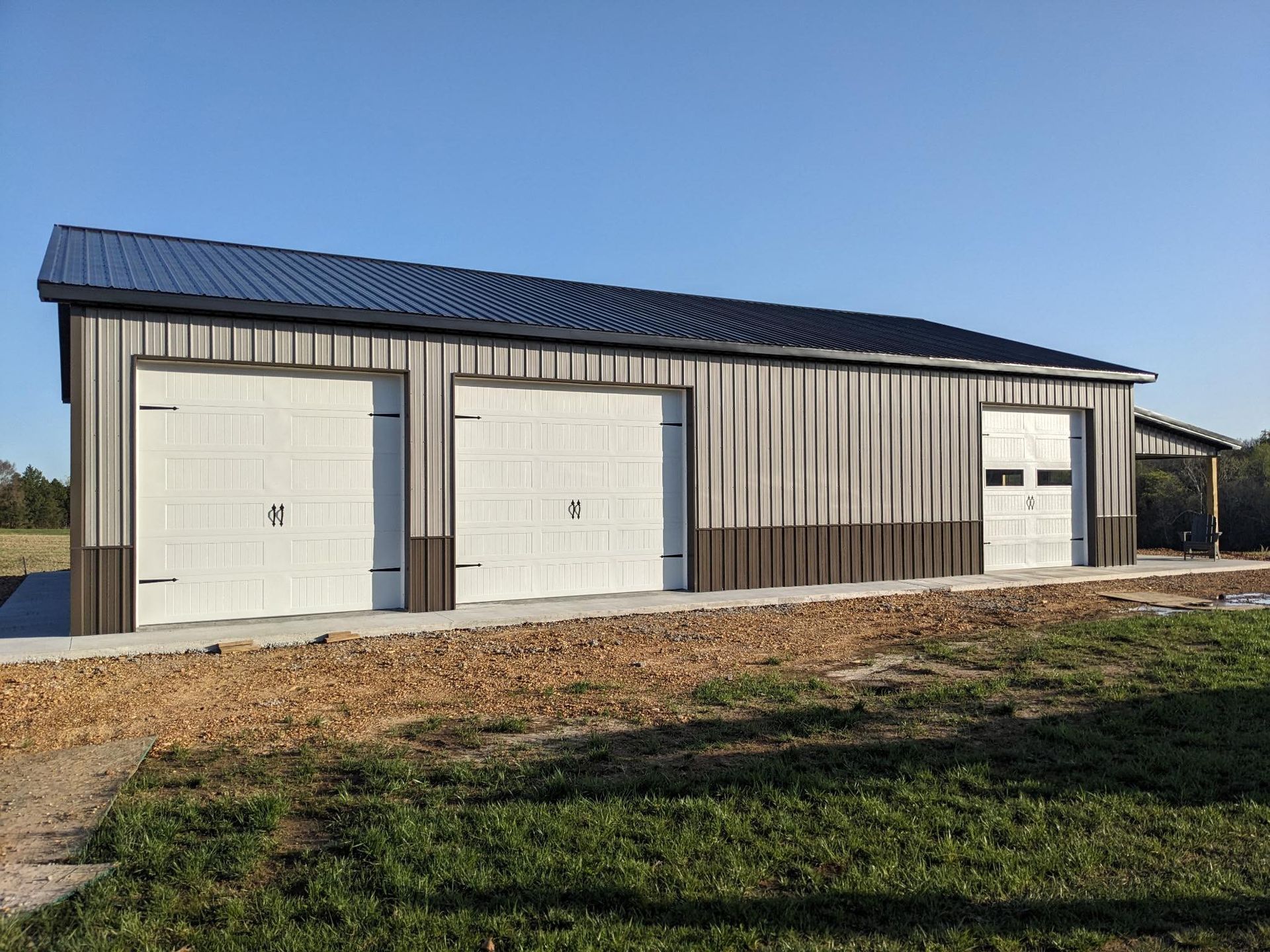 Metal garage with three doors, gray and brown siding, dark roof, set on concrete with grass.