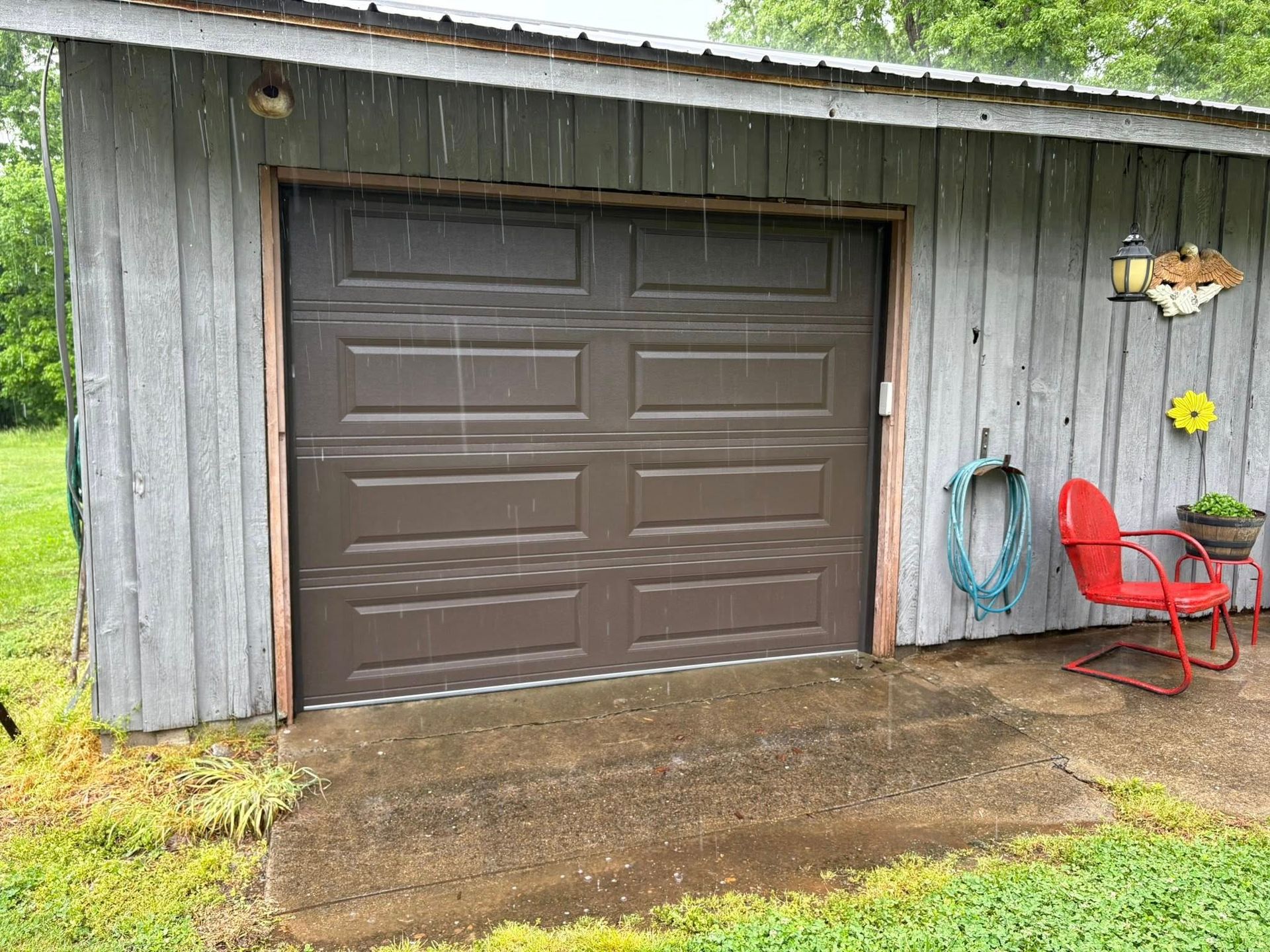 Brown garage door on a gray weathered shed during rain. Red chair and hose visible.