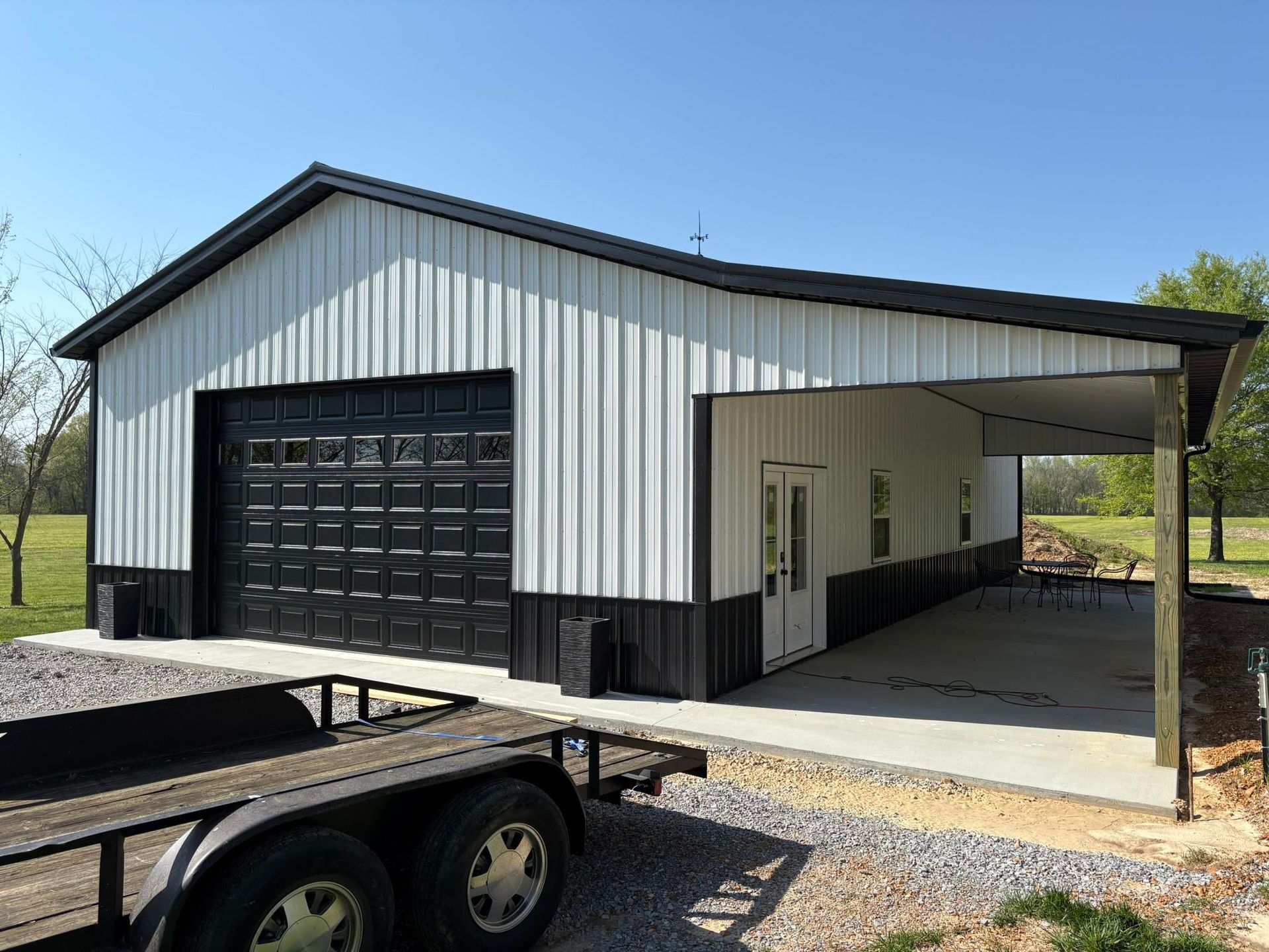 Metal building with black and white siding, garage door, and carport. A trailer is parked in front.