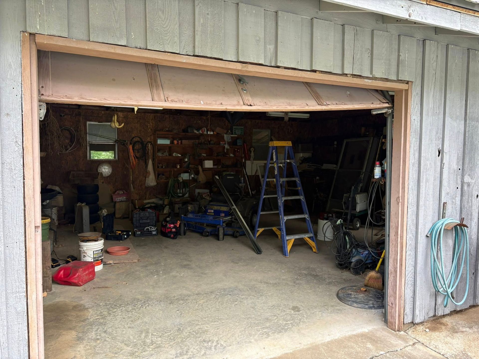 Garage interior, cluttered with tools and equipment. Garage door partially open, revealing a messy space.