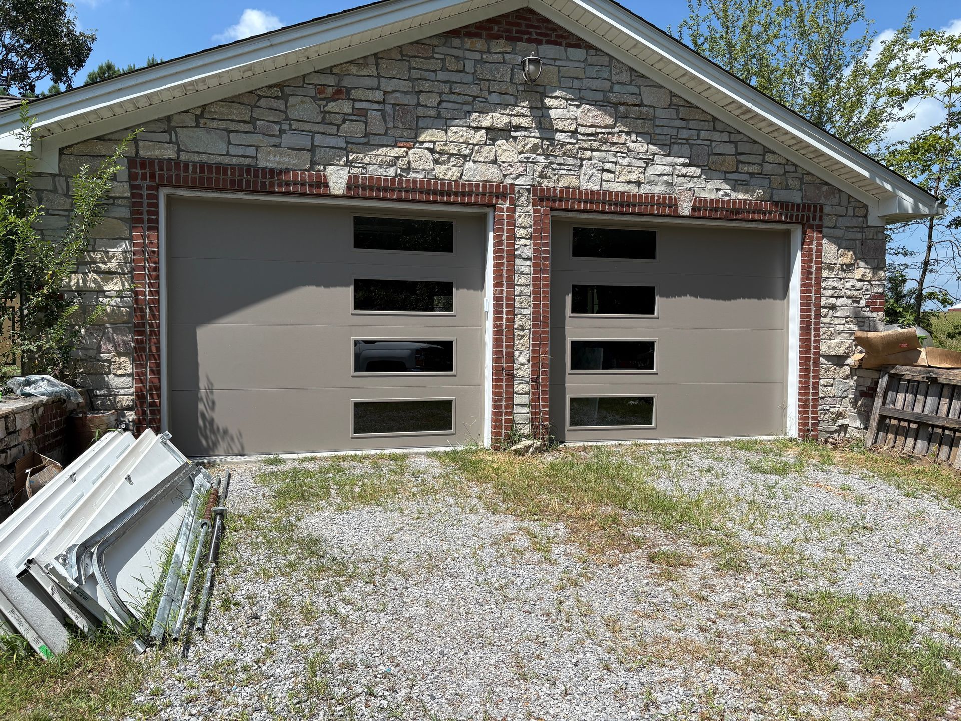 Two tan garage doors with rectangular windows on a stone building, on a gravel driveway.