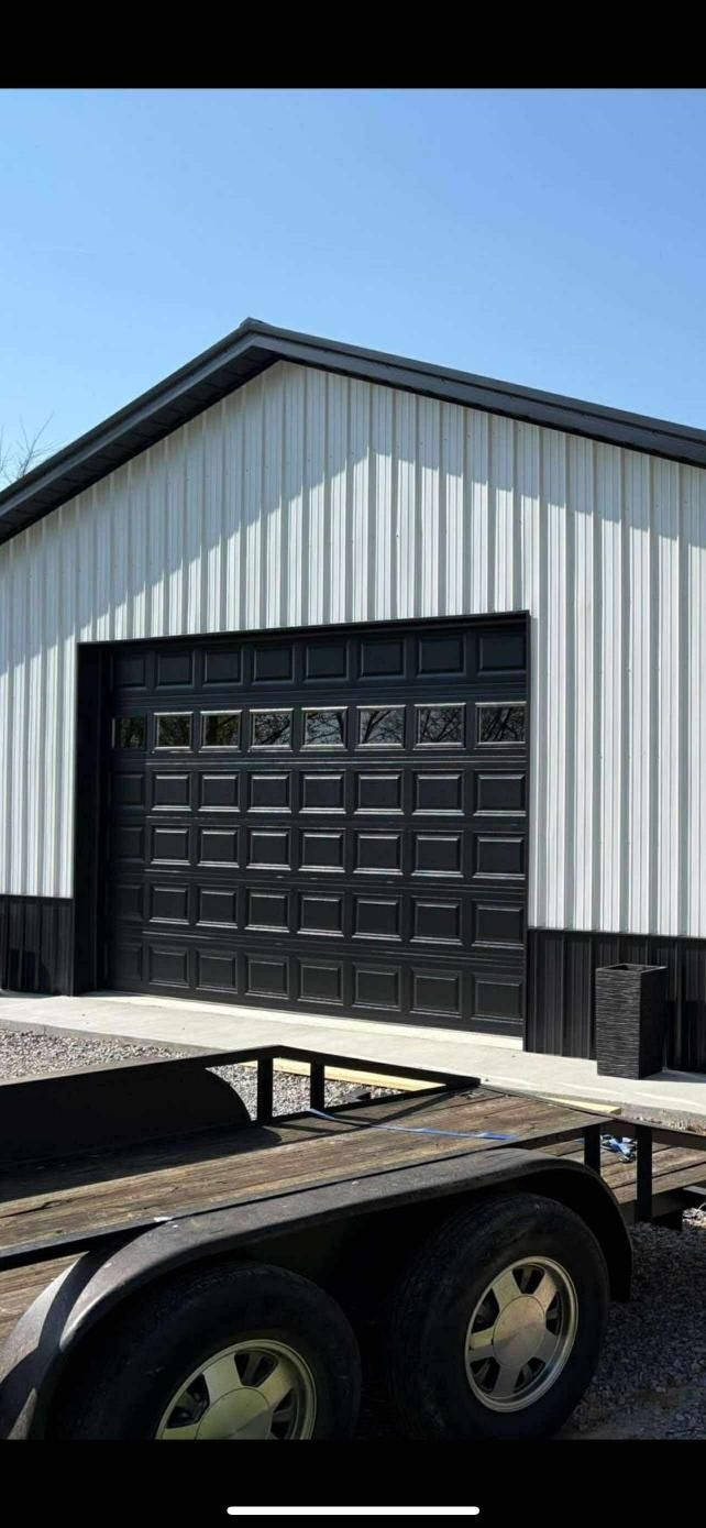 White and black metal building with a black garage door. Trailer in foreground, blue sky.