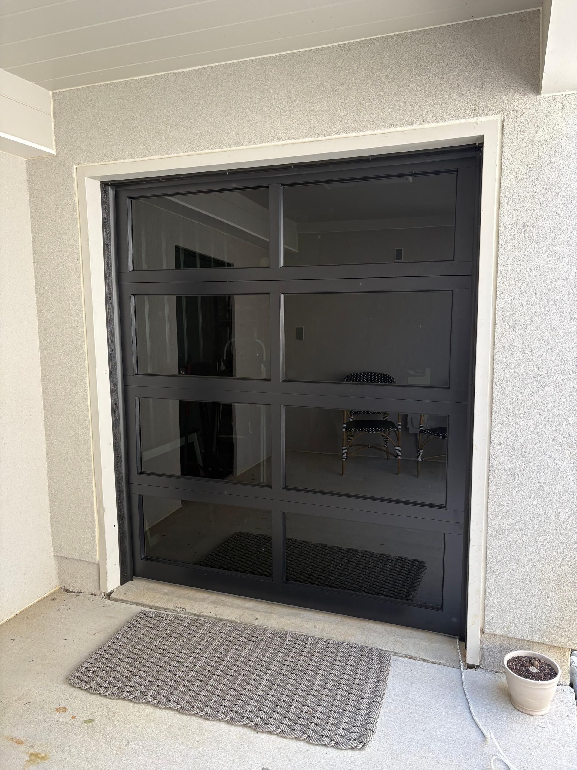 Black glass door with grid pattern, gray door mat, and small potted plant in an outdoor setting.