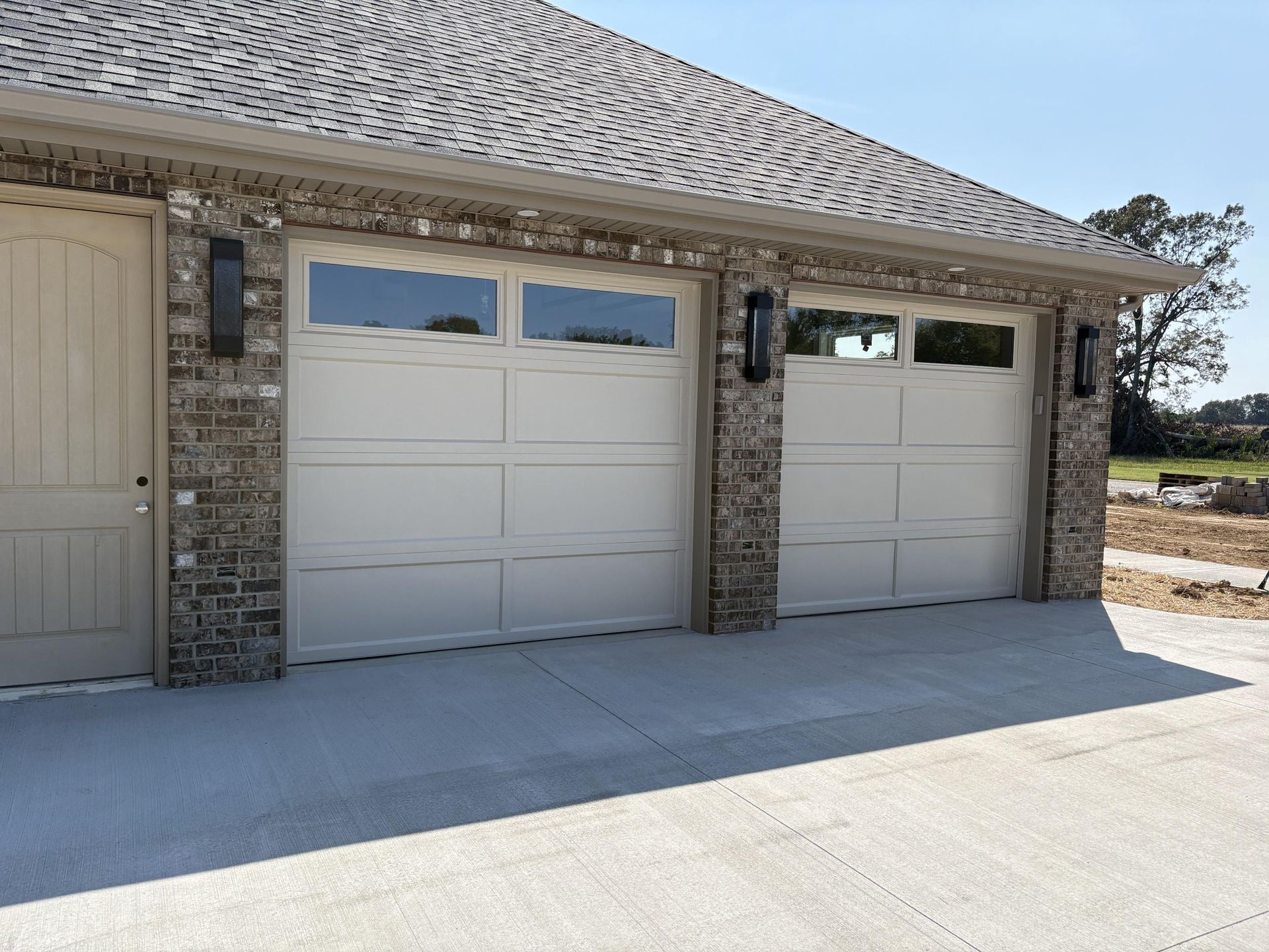 Two tan garage doors on a brick building with a concrete driveway. Black light fixtures.