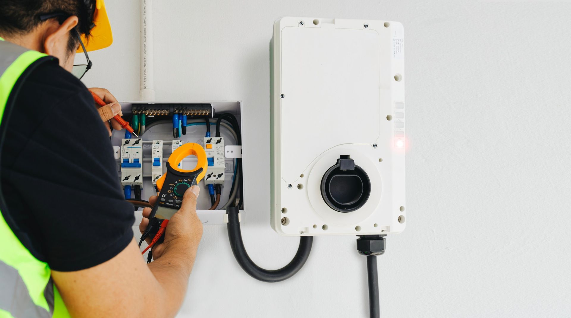 Electrician in safety vest using multimeter to inspect electrical panel next to EV charger.
